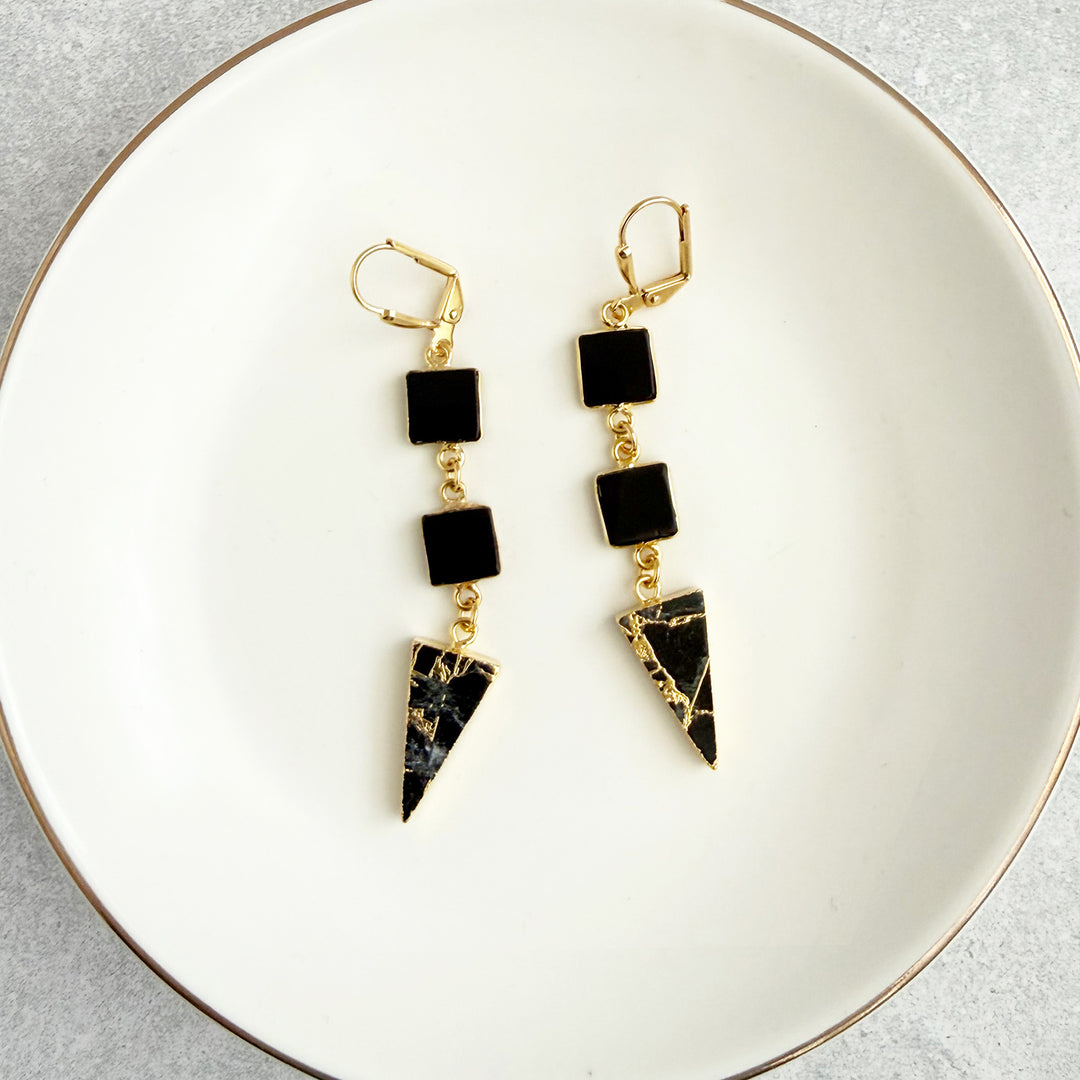 Pair of gold earrings with black square and triangle-shaped stones on a white plate.